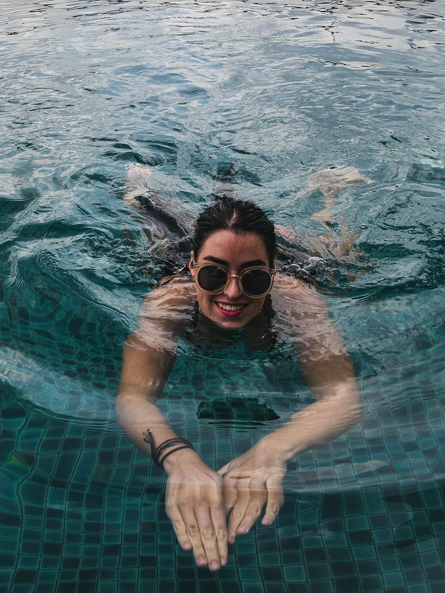 Smiling person with sunglasses swimming towards camera in tiled pool.