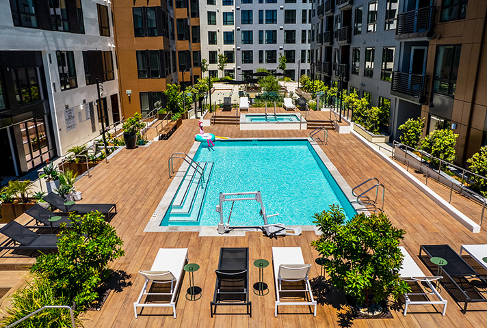 Pool deck with lounge chairs, umbrellas, and hot tub overlooked by apartments.